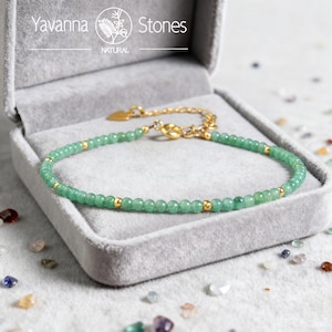 Könnte beinhalten: Ein zartes Armband mit grünen Perlen und goldenen Akzenten, präsentiert in einer grauen Samt-Schmuckschatulle. Das Armband hat kleine, runde grüne Perlen, die mit goldfarbenen Perlen durchsetzt sind. Der Verschluss und die Kette sind ebenfalls goldfarben. Der Markenname "Yavanna Stones" ist sichtbar.