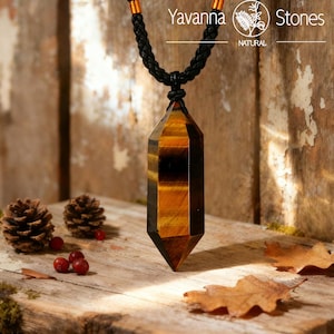 May include: A faceted tiger's eye pendant necklace with a black braided cord. The gemstone is a mix of brown and gold tones. The necklace is displayed on a rustic wooden surface with pinecones, berries, and leaves. The brand name "Yavanna Stones" is visible.
