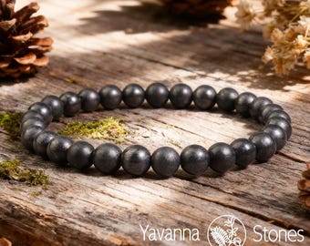 May include: A dark gray beaded bracelet with round beads. The bracelet is displayed on a rustic wooden surface with pine cones and moss. The Yavanna Stones logo is visible in the bottom right corner.
