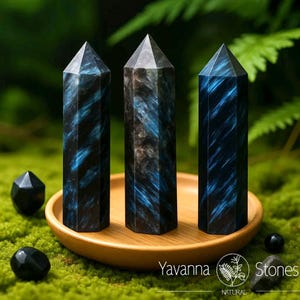 May include: Three dark blue and black crystal obelisks with pointed tops are displayed on a wooden tray, surrounded by small black stones and greenery. The obelisks have a striking blue veining pattern. The tray sits on a bed of green moss.