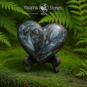 May include: A heart-shaped stone with blue and brown veining, displayed on a small black stand. The stone is set against a backdrop of green moss and fern leaves. The text "Yavanna Stones NATURAL" is visible.