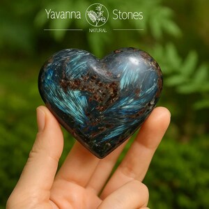 May include: A heart-shaped stone with a mix of blue, brown, and black colors. The stone is held in a hand, with the background blurred. The top of the image has the text "Yavanna Stones NATURAL".