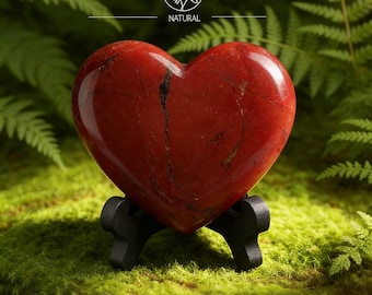 Puede incluir: Una piedra en forma de corazón de jaspe rojo pulido, exhibida sobre un soporte negro. La piedra tiene vetas oscuras y está sobre musgo verde, con helechos en el fondo. El texto "Yavanna Stones NATURAL" es visible.