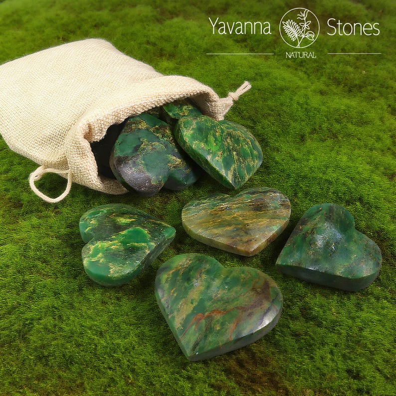 May include: A collection of heart-shaped green stones, some with brown and gold veining, spilling from a beige drawstring bag. The stones are arranged on a bed of green moss. The image includes the text "Yavanna Stones NATURAL".