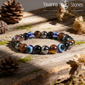 May include: A beaded bracelet featuring a variety of stone beads. The bracelet includes brown, black, and blue beads, some with an eye design. The bracelet is displayed on a wooden surface with pine cones and moss.