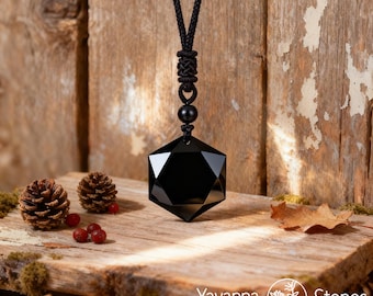 Puede incluir: Un collar con un colgante geométrico negro y un cordón anudado. El colgante tiene una forma multifacética que refleja la luz. El collar cuelga sobre una superficie de madera con piñas y bayas. El texto "Yavanna Stones" es visible.