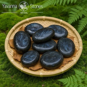 May include: A selection of polished, oval stones with shimmering blue and grey patterns, presented in a woven bamboo basket. The stones rest on brown paper, set against a backdrop of green moss and foliage. The image includes the text "Yavanna Stones NATURAL".