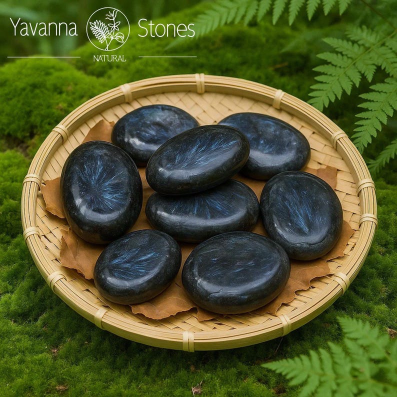 May include: A selection of polished, oval stones with shimmering blue and grey patterns, presented in a woven bamboo basket. The stones rest on brown paper, set against a backdrop of green moss and foliage. The image includes the text "Yavanna Stones NATURAL".