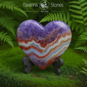 May include: A heart-shaped stone sculpture with a layered design in shades of purple, red, orange, and white. The stone is displayed on a dark gray stand against a backdrop of green foliage. The text "Yavanna Stones NATURAL" is visible.