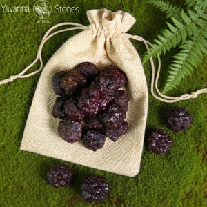 May include: A beige drawstring bag filled with dark purple, textured stones. The bag is on green moss, with some stones scattered around. The image has the text "Yavanna Stones NATURAL" in the top left corner.