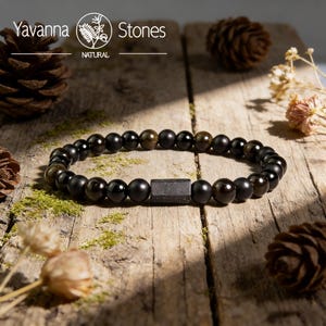 May include: Black beaded bracelet with a rectangular accent. The bracelet is made of round, dark beads with a few golden-brown beads. The bracelet is displayed on a rustic wooden surface with pine cones and dried flowers. The text "Yavanna Stones NATURAL" is at the top.