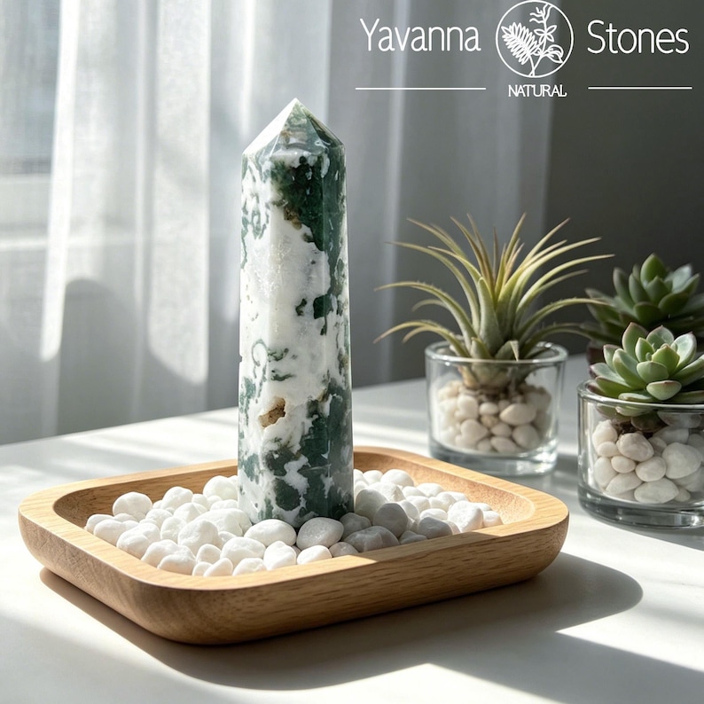 May include: A polished moss agate crystal tower with white and deep green inclusions, standing upright in a rectangular wooden tray filled with small white stones. The text Yavanna Stones Natural is visible in the upper right corner of the image.