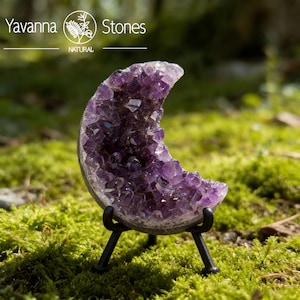 Pode incluir: Uma geodo de ametista em forma de crescente, exibindo um tom roxo profundo, é exibida em um pequeno suporte de metal preto. A estrutura cristalina da geodo é visível, com um fundo de musgo verde. O texto "Yavanna Stones NATURAL" também é visível.