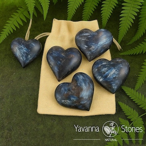 May include: A collection of heart-shaped stones in shades of blue and black, some with a white marbling effect. One heart is suspended by a string. The stones are displayed with a beige drawstring bag and green foliage.