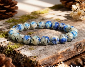 May include: A beaded bracelet made of round, polished k2 stone beads. The beads are a mix of white, gray, and blue, with a speckled pattern. The bracelet is displayed on a piece of wood with moss and pine cones.