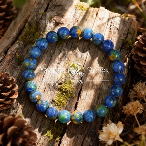 May include: A beaded bracelet featuring round, blue and green stones with yellow accents. The bracelet is displayed on a weathered wooden surface with moss and pine cones. The text "Yavanna Stones NATURAL" is visible.