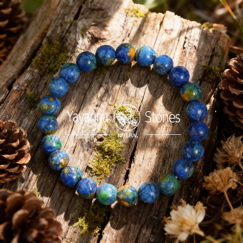 May include: A beaded bracelet featuring round, blue and green stones with yellow accents. The bracelet is displayed on a weathered wooden surface with moss and pine cones. The text "Yavanna Stones NATURAL" is visible.