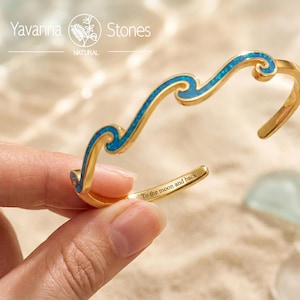 May include: A gold-toned cuff bracelet featuring a wave-shaped design inlaid with shimmering blue opal-like material. The inner surface of the metal band is engraved with the text To the moon and back in a clear, dark, sans-serif font.