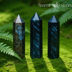 May include: Three natural, polished crystal obelisks with pointed tops. The obelisks are dark blue and brown with streaks of lighter blue. The crystals are displayed on a bed of green moss. The text "Yavanna Stones NATURAL" is at the top.