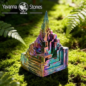 May include: A vibrant, multi-colored bismuth crystal sculpture with a layered, geometric design. The crystal displays iridescent hues of blue, purple, gold, and green. The sculpture is set against a backdrop of green moss and ferns. The text "Yavanna Stones NATURAL" is at the top.