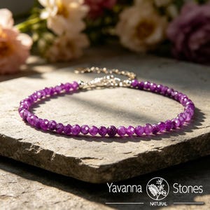 May include: A delicate bracelet featuring small, faceted, deep purple gemstones strung together. The bracelet has a silver-colored clasp and chain. The jewelry is displayed on a gray stone surface, with blurred flowers in the background.