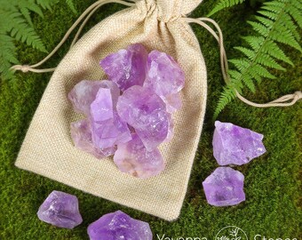May include: A collection of raw, natural amethyst crystals in varying shades of purple, spilling from a beige drawstring bag. The crystals are scattered on a bed of green moss with fern leaves. The Yavanna Stones logo is visible.
