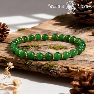 May include: A green jade bead bracelet with gold-colored accents. The bracelet is displayed on a piece of wood with moss and pine cones. The brand name "Yavanna Stones" is visible in the top right corner.