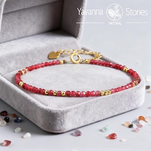 Puede incluir: Una delicada pulsera con pequeñas cuentas rojas facetadas intercaladas con cuentas doradas. La pulsera se muestra en una caja de joyería gris. El cierre y la cadena son de color dorado. La marca "Yavanna Stones" es visible.