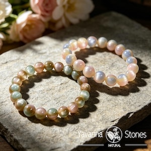 May include: Two elastic bracelets composed of smooth, round, iridescent beads. One bracelet features soft pink and pale blue tones, while the other displays muted green, gold, and peach hues. The text Yavanna Stones Natural is visible.