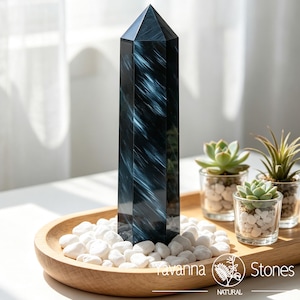 May include: A tall, polished, hexagonal crystal tower featuring deep navy blue and black tones with striking, light blue fibrous streaks. The crystal stands upright among white decorative stones on a wooden tray with small succulents.