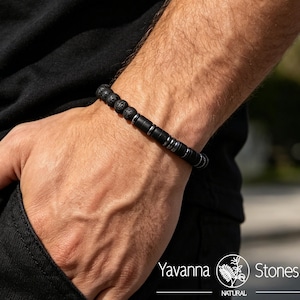 May include: A beaded bracelet featuring a combination of textured black lava stone beads and smooth, cylindrical black metal spacers. The bracelet is worn on a wrist and includes the text Yavanna Stones Natural in the bottom corner.