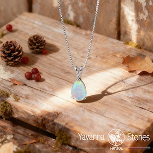 May include: A silver necklace with a pear-shaped opal pendant. The opal displays a mix of colors, including blue, green, and yellow. The necklace is displayed on a rustic wooden surface with pinecones and berries.