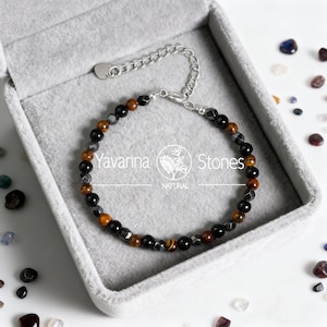May include: A beaded bracelet with a silver chain clasp, presented in a gray jewelry box. The bracelet features a mix of black, brown, and gray beads, including tiger's eye stones. The brand name "Yavanna Stones" is visible.