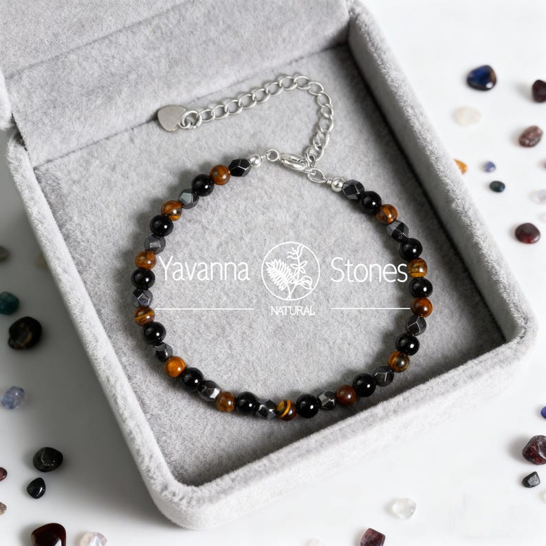 May include: A beaded bracelet with a silver chain clasp, presented in a gray jewelry box. The bracelet features a mix of black, brown, and gray beads, including tiger's eye stones. The brand name "Yavanna Stones" is visible.