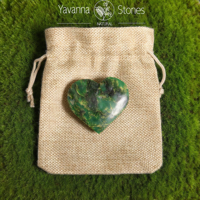 May include: A heart-shaped green stone with dark green and gold veining rests on a beige burlap bag. The bag is closed with a drawstring. The words "Yavanna Stones NATURAL" are printed at the top.
