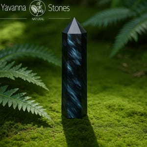 Op de afbeelding: Een donkerblauwe kristallen obelisk met een puntige top, geplaatst tegen groene mos en varens. Het kristal heeft een uniek patroon van lichtblauwe strepen. De tekst "Yavanna Stones" staat in de linkerbovenhoek.