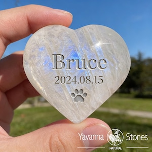 May include: Heart-shaped, polished stone with the name "Bruce", date "2024.08.15", and a paw print engraved on the surface. The stone has a pearly white color with blue and iridescent highlights. The Yavanna Stones logo is visible.
