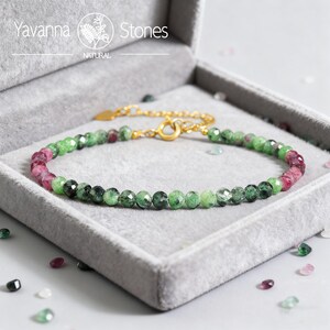 May include: A beaded bracelet with green and pink gemstones, displayed in a gray velvet box. The bracelet has a gold-colored chain and clasp. The brand name "Yavanna Stones" is visible in the top left corner.