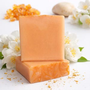 May include: Two stacked, square-shaped bars of orange soap. The top bar is smooth, while the bottom bar has a swirled design. White jasmine flowers and golden granules surround the soap, creating a natural, spa-like aesthetic.