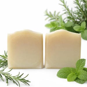 May include: Two square, light-colored soaps are displayed, surrounded by sprigs of fresh rosemary and mint leaves. The soaps have a slightly uneven top edge, suggesting a handmade quality. The overall aesthetic is clean and natural.