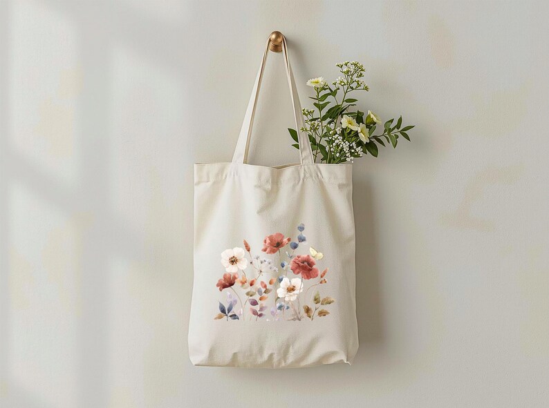 May include: A cream-colored tote bag with a floral design of red, white, and blue flowers. The bag is hanging on a gold hook, and a bouquet of white and yellow flowers is inside the bag.