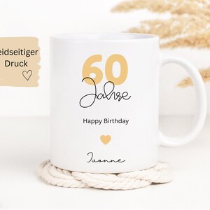 May include: White ceramic mug with a gold "60 Jahre" design, "Happy Birthday", and a name. The mug sits on a white rope coaster. The mug has a handle and is decorated with a gold heart.