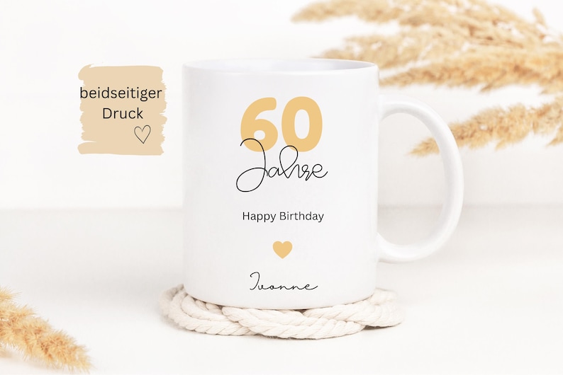 May include: White ceramic mug with a gold "60 Jahre" design, "Happy Birthday", and a name. The mug sits on a white rope coaster. The mug has a handle and is decorated with a gold heart.
