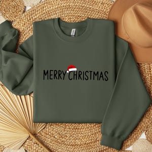 May include: An olive green crewneck sweatshirt featuring the black text MERRY CHRISTMAS printed across the chest. A small red and white Santa hat graphic is positioned above the letter R in the word MERRY.