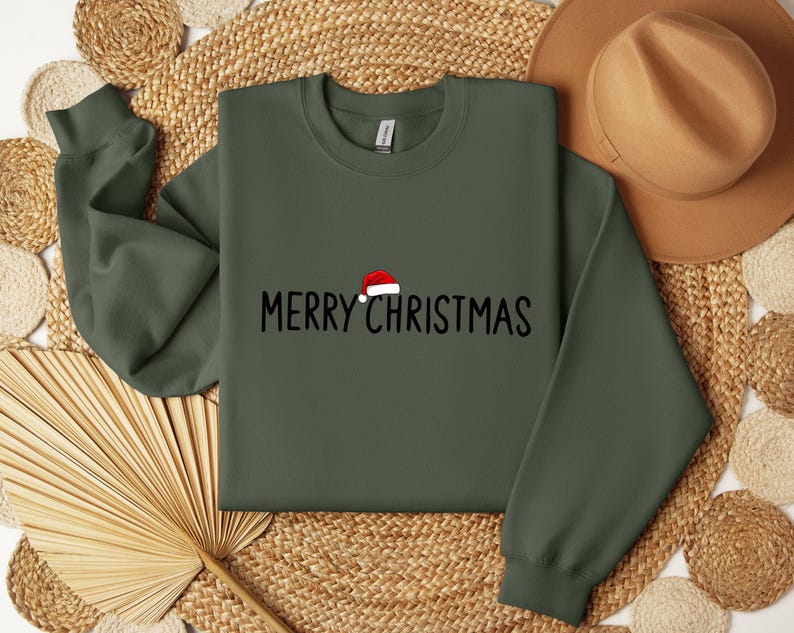 May include: An olive green crewneck sweatshirt featuring the black text MERRY CHRISTMAS printed across the chest. A small red and white Santa hat graphic is positioned above the letter R in the word MERRY.