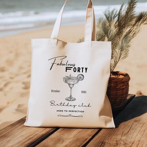 May include: A cream-coloured tote bag featuring the words "Fabulous Forty" and a cocktail illustration. Below the illustration are the words "October 1985" and "Birthday Club". The bag is on a wooden surface, with a beach scene in the background.