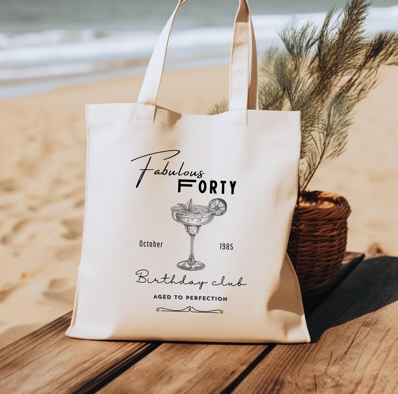 May include: A cream-coloured tote bag featuring the words "Fabulous Forty" and a cocktail illustration. Below the illustration are the words "October 1985" and "Birthday Club". The bag is on a wooden surface, with a beach scene in the background.