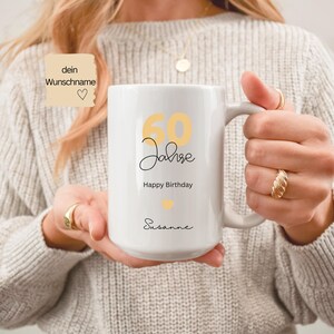 May include: White ceramic mug with a handle, featuring the text "60 Jahre Happy Birthday Susanne" in black and yellow. The mug has a heart symbol and is held by a person.