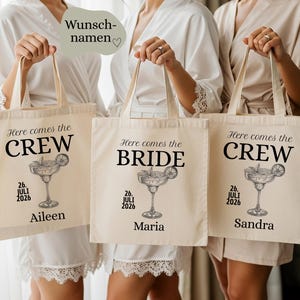 May include: Three cream-colored tote bags with black text and a margarita illustration. The text on the bags reads "Here comes the CREW", "Here comes the BRIDE", and "Here comes the CREW". Each bag has a name and the date "26. JULI 2026".