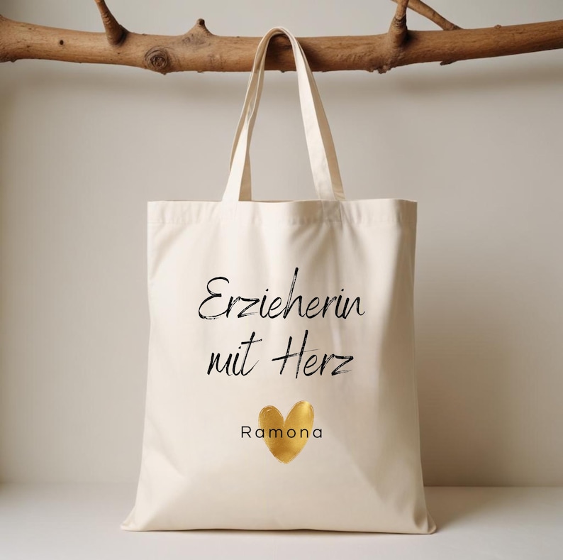 May include: Cream-colored tote bag with the text "Erzieherin mit Herz" and the name "Ramona" below a gold heart. The bag has two handles and is hanging in front of a light background.